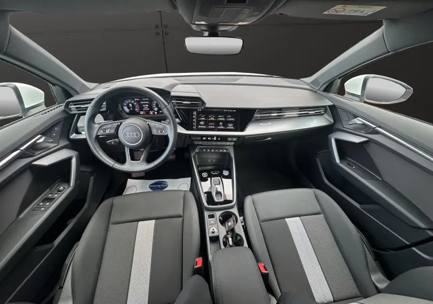 Intérieur noir de l'Audi A3 Sportback 2024 vu de face, avec tableau de bord digital et sièges tissu à bande grise centrale.