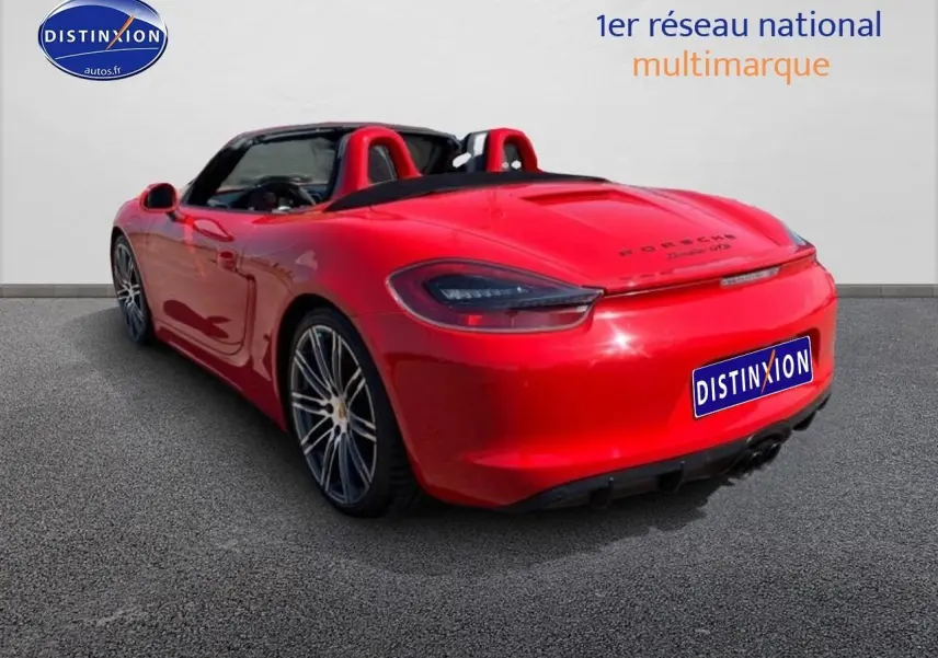 Vue 3/4 arrière droite d'une Porsche Boxster 981 rouge avec capote ouverte et jantes alu multibranches.