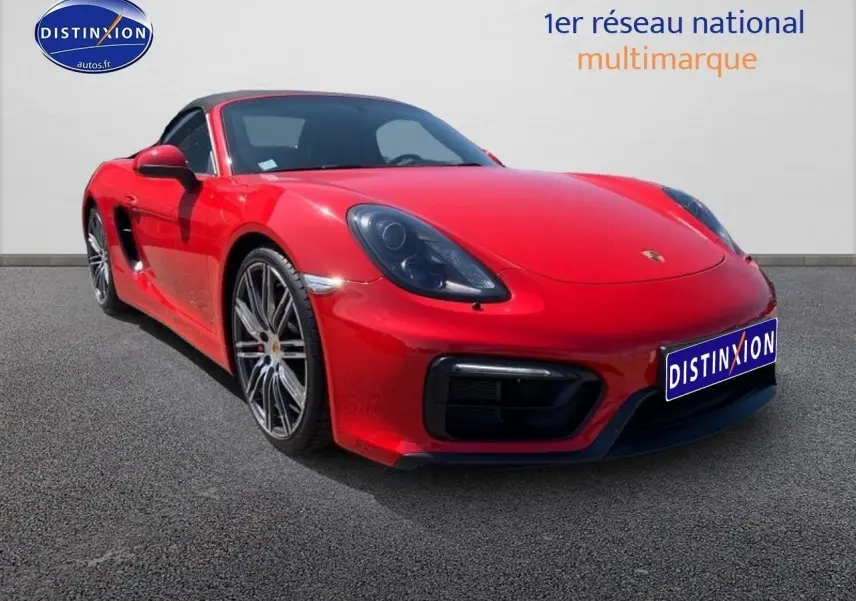 Porsche Boxster rouge vue 3/4 avant droit avec jantes alu et capote souple noire sur sol gris.