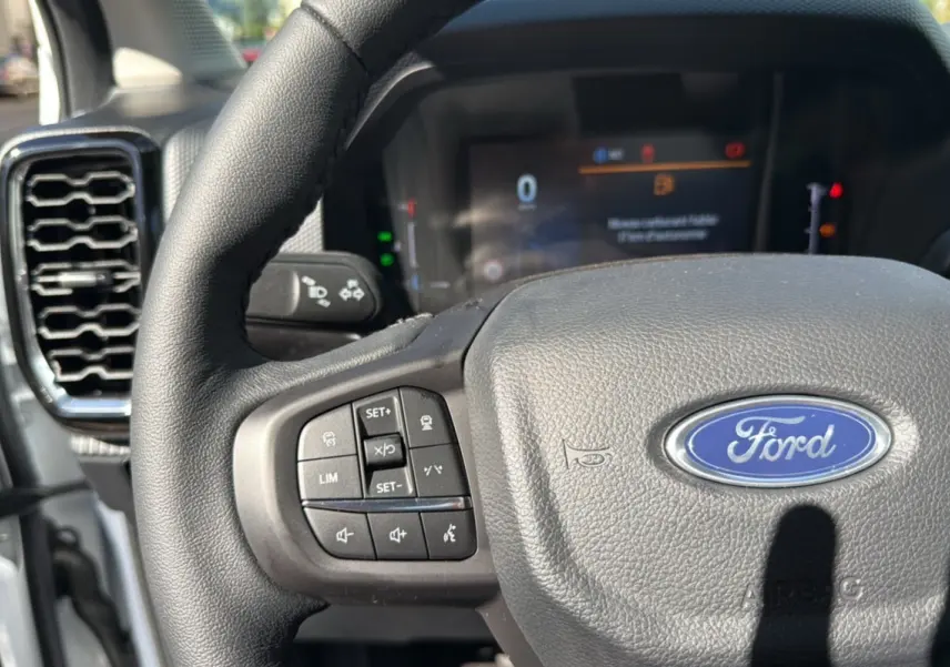 Vue rapprochée du volant cuir noir du Ford Ranger blanc glacier avec commandes multifonctions et tableau de bord numérique flou.