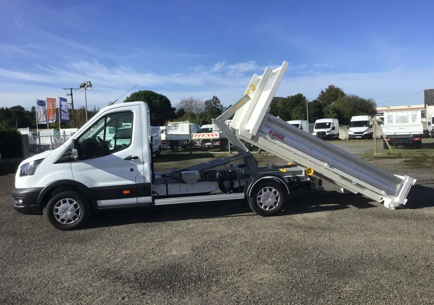 Vue latérale gauche d'un Ford Transit blanc avec benne relevée et bras polybenne sur un terrain extérieur.