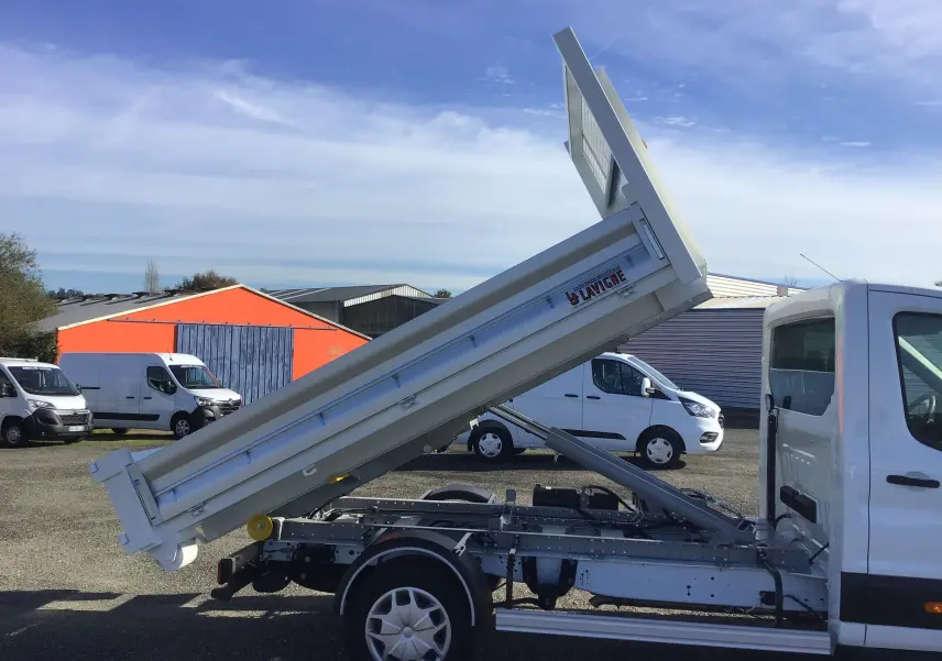 Vue latérale droite d'un Ford Transit blanc avec benne relevée et bras polybenne visible sur un parking extérieur.