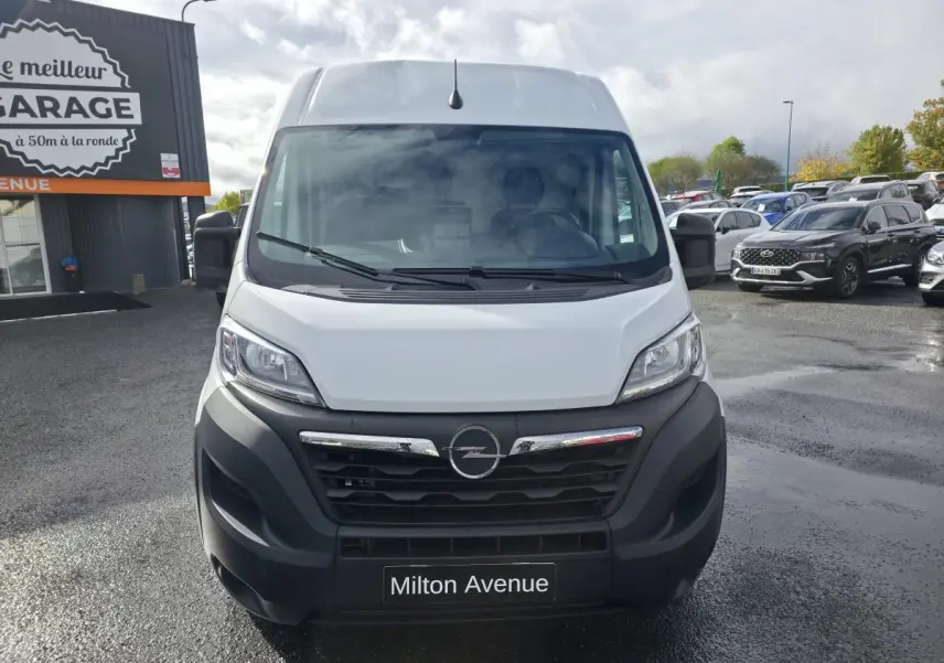Vue de face d'un fourgon Opel Movano blanc avec calandre noire et logo Opel au centre, stationné devant un garage.
