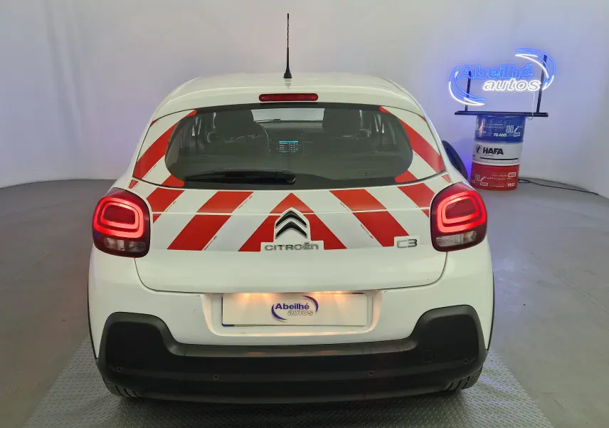 Vue arrière d'une Citroën C3 blanche 2019 avec bandes rouges diagonales de signalisation et feux arrière allumés.