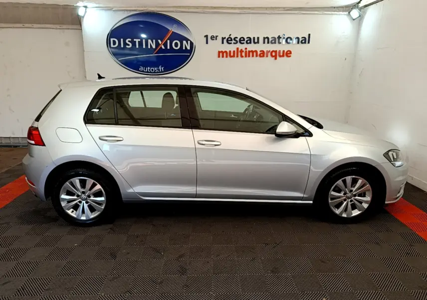 Profil côté gauche d'une Volkswagen Golf gris clair 2019 garée en intérieur avec logo Distinxion en arrière-plan.