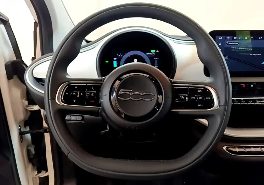 Vue intérieure centrée sur le volant noir de la Fiat 500 électrique blanche avec tableau de bord digital et écran tactile.