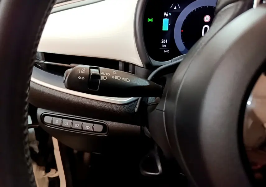 Gros plan sur le combiné d'instruments numérique et la commande des phares à gauche du volant dans le tableau de bord blanc de la Fiat 500 électrique blanche.