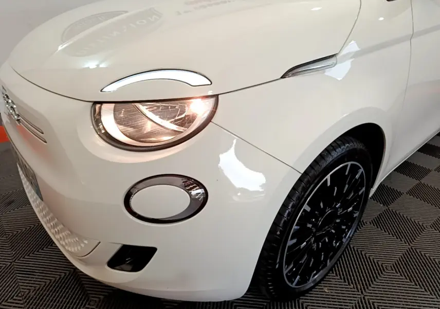 Vue 3/4 avant droit d'une Fiat 500 électrique blanche avec phare allumé et jante noire design moderne.