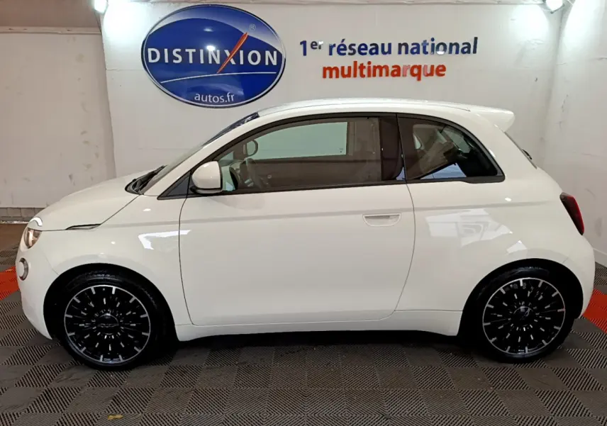 Profil côté gauche d'une FIAT 500 électrique blanche 42kWh Icône 2022 avec jantes noires stylisées en intérieur showroom.