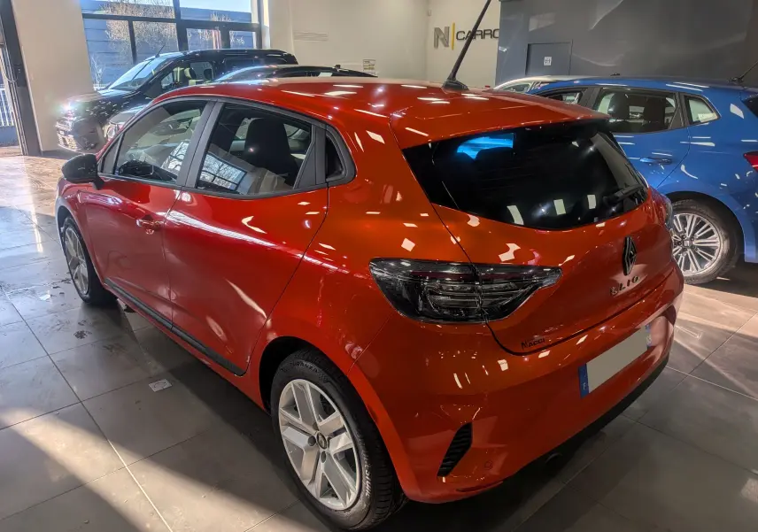 Vue 3/4 arrière droite d'une Renault Clio V Orange Valencia 2024 en intérieur, avec feux arrière fumés et antenne sur le toit.