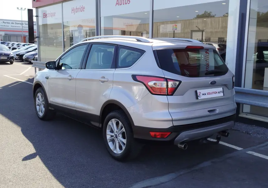 Vue 3/4 arrière droite d'un Ford Kuga gris clair 2019 avec barres de toit et double sortie d'échappement visible.