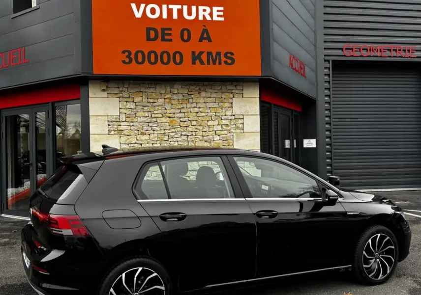 Volkswagen Golf noire vue de profil côté gauche, stationnée devant un bâtiment avec panneau orange "Votre nouvelle voiture".