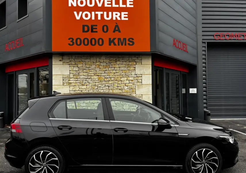 Profil côté gauche d'une Volkswagen Golf noire 2024 garée devant un bâtiment avec panneau orange "Votre nouvelle voiture".