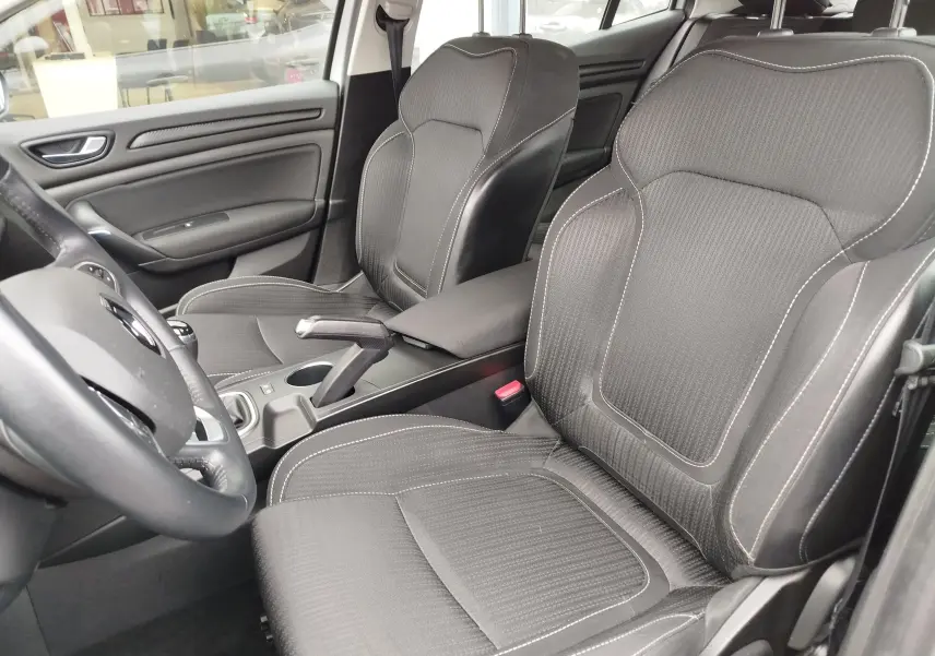 Vue intérieure côté conducteur de la Renault Mégane 2018, sièges noirs avec surpiqûres blanches et volant partiellement visible.