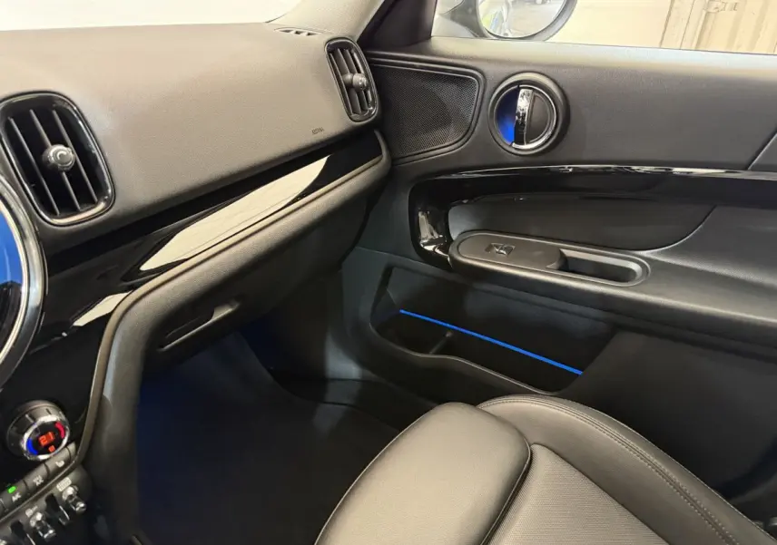 Vue intérieure côté passager de la MINI Countryman gris clair 2023, tableau de bord et porte avec éclairage d'ambiance bleu.
