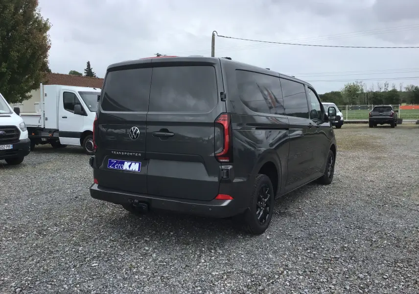Vue 3/4 arrière droite d'un Volkswagen Transporter T7 gris anthracite avec barres de toit et jantes noires sur parking gravel.