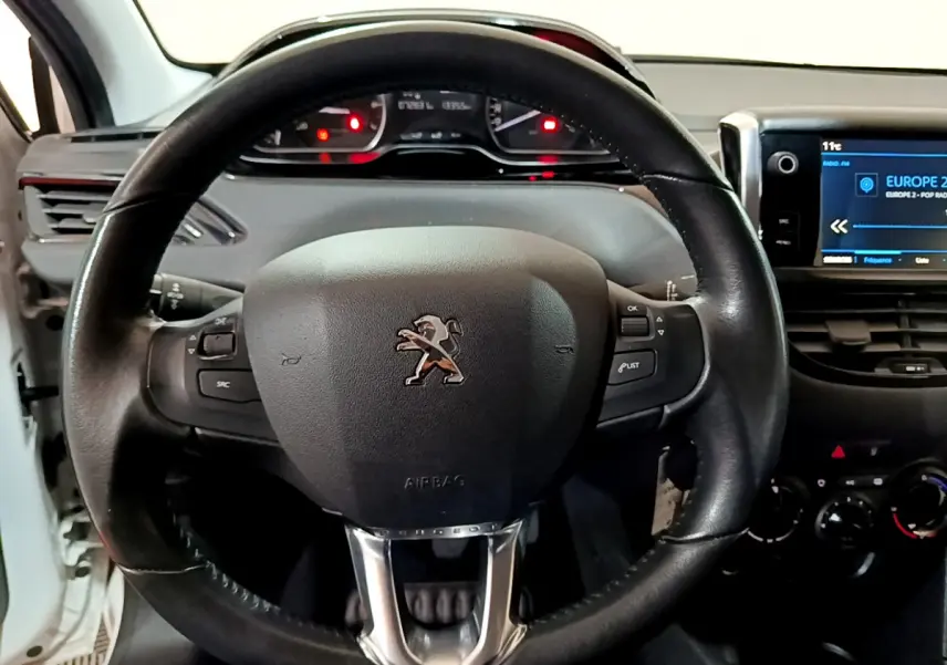 Vue rapprochée du volant cuir noir avec logo Peugeot et tableau de bord numérique d'une 208 blanche de 2018.