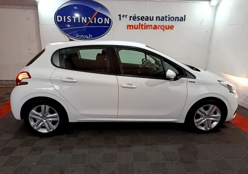 Profil côté gauche d'une Peugeot 208 Puretech 82 Signature blanche, en intérieur sur sol damier avec feux arrière allumés.