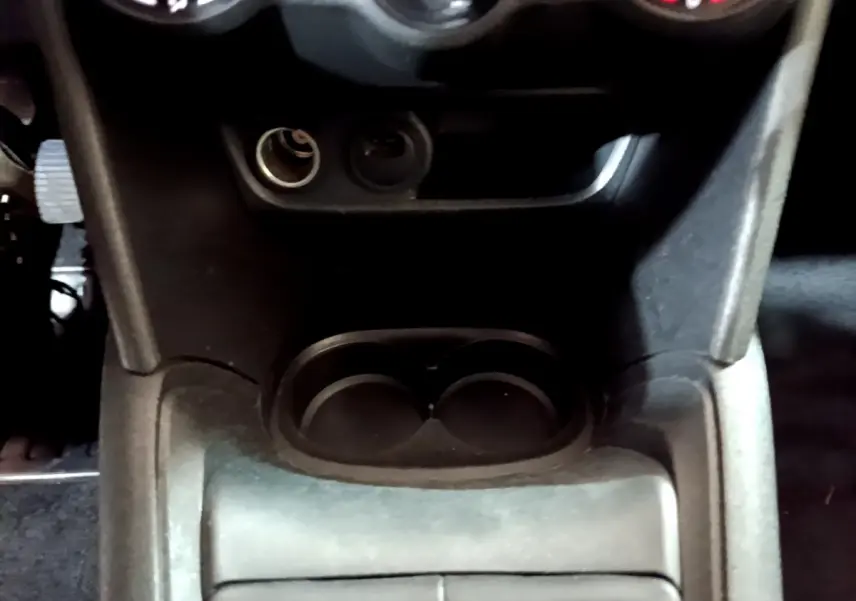 Gros plan sur la console centrale noire de la Peugeot 208 Puretech 82 Signature, avec porte-gobelets et prise 12V visible.