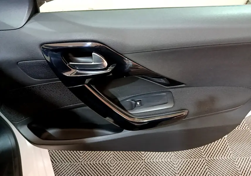 Gros plan sur la porte intérieure côté conducteur d'une Peugeot 208 blanche, avec poignée noire brillante et commande de vitre.