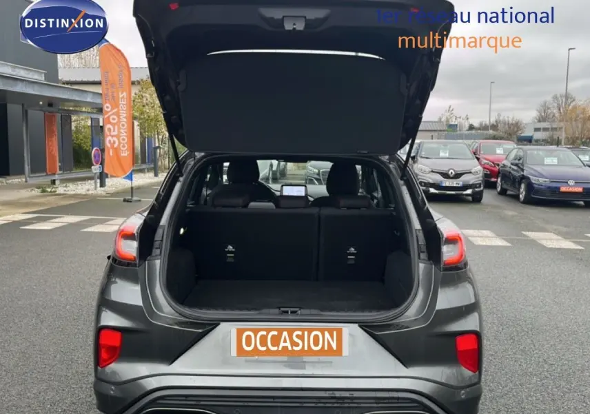 Vue arrière d'un Ford Puma gris Magnetic Metal 2024 avec coffre ouvert sur un parking de voitures d'occasion.