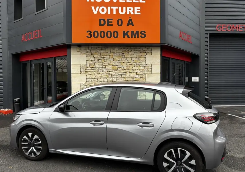 Peugeot 208 PureTech 100 S&S Active gris clair vue de profil côté gauche devant un bâtiment avec panneau orange "Votre nouvelle voiture".