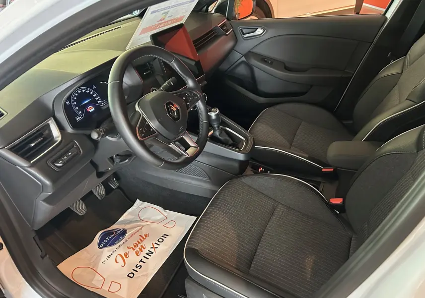 Vue intérieure côté conducteur de la Renault Clio V blanc nacré, montrant le tableau de bord et les sièges noirs.