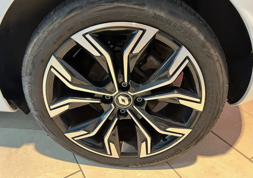 Gros plan sur la jante noire et argentée d'une Renault Clio V blanc nacré, mettant en valeur le logo central Renault.