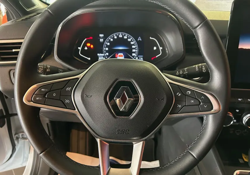 Vue rapprochée du volant en cuir noir avec logo Renault et tableau de bord numérique éclairé d'une Clio V blanche nacrée.