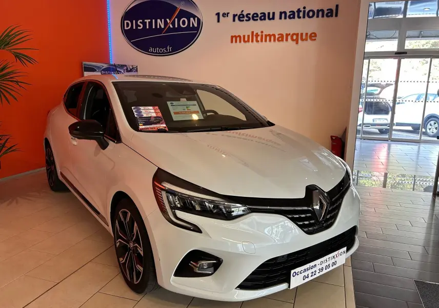 Renault Clio V blanc nacré métal vue 3/4 avant droit en showroom avec jantes noires et stickers vitres.