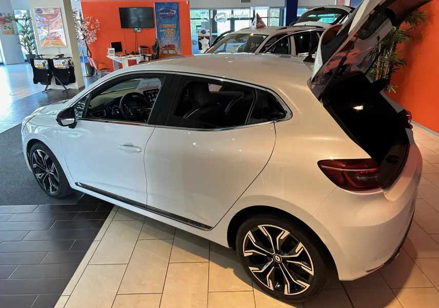 Renault Clio V blanc nacré vue de côté droit arrière avec coffre ouvert en showroom lumineux.