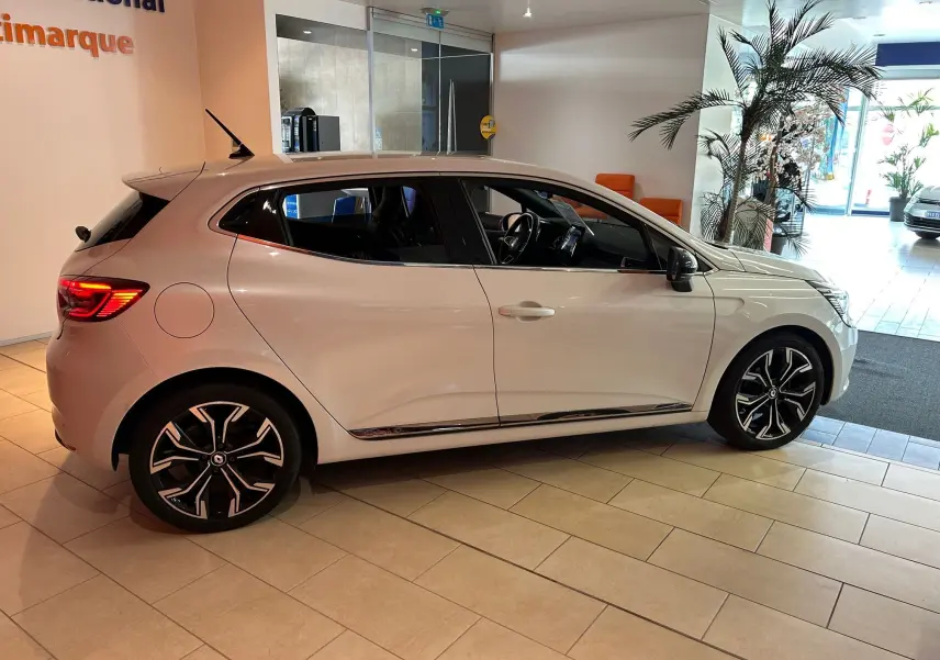 Profil droit d'une Renault Clio V blanche nacrée avec jantes alliage noires et détails chromés en intérieur showroom.