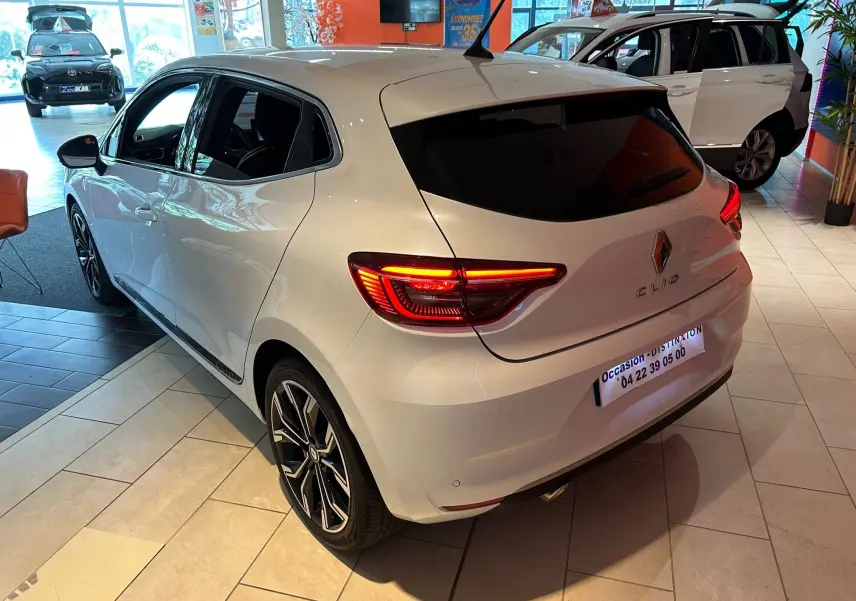 Vue 3/4 arrière droite d'une Renault Clio V blanc nacré métal avec feux arrière allumés en showroom.