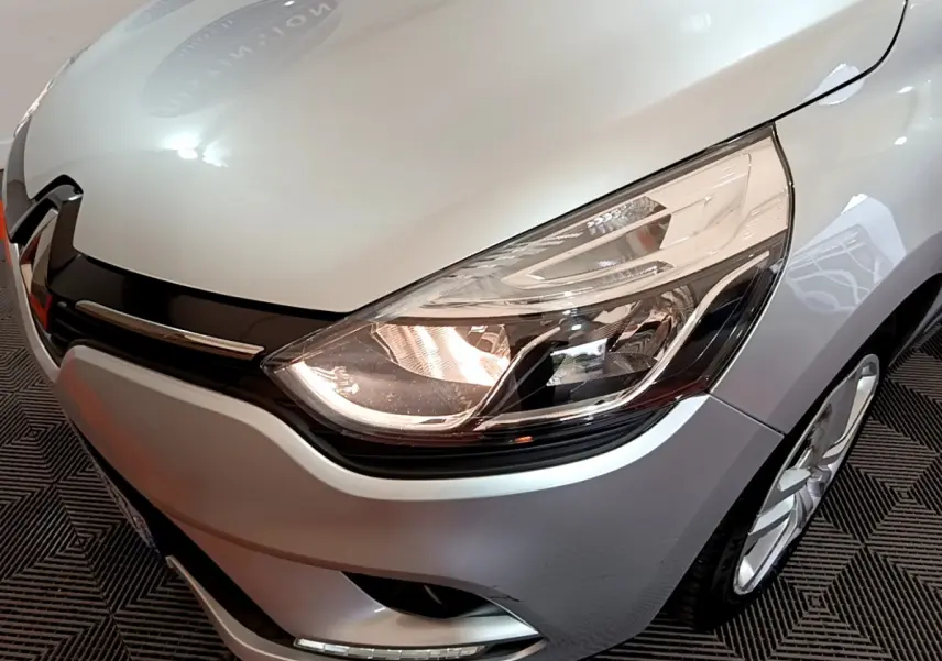 Gros plan sur l'avant droit d'une Renault Clio Business TCe 90 gris clair, mettant en valeur le phare allumé et la calandre noire.
