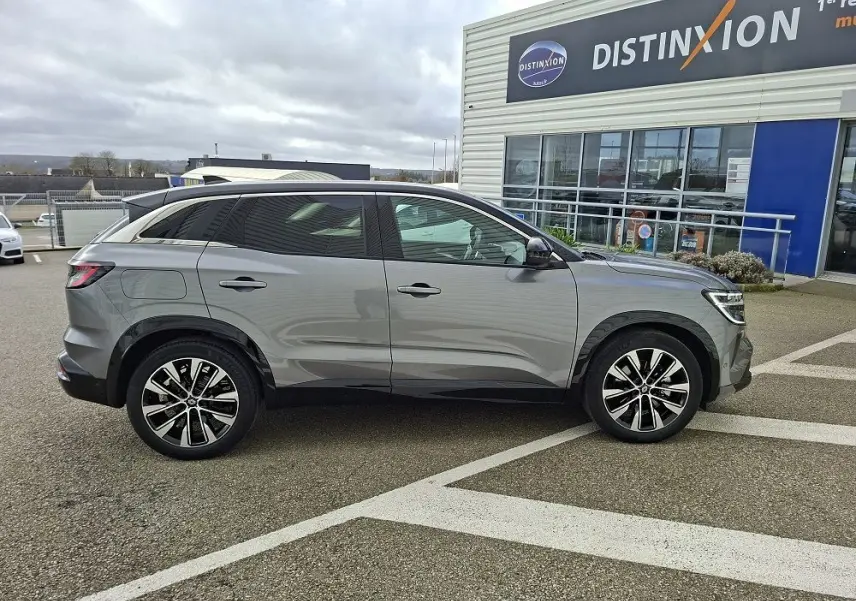 Profil droit d’un Renault Austral E-Tech Full Hybrid gris schiste, avec jantes alliage et vitres teintées, stationné devant un bâtiment.