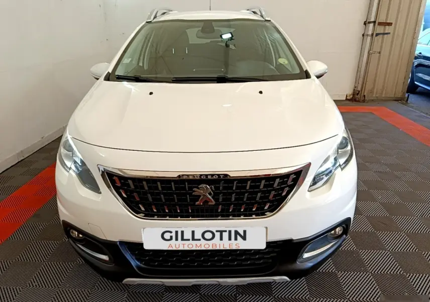 Vue avant d'un Peugeot 2008 blanc avec calandre chromée et phares allumés en intérieur showroom.