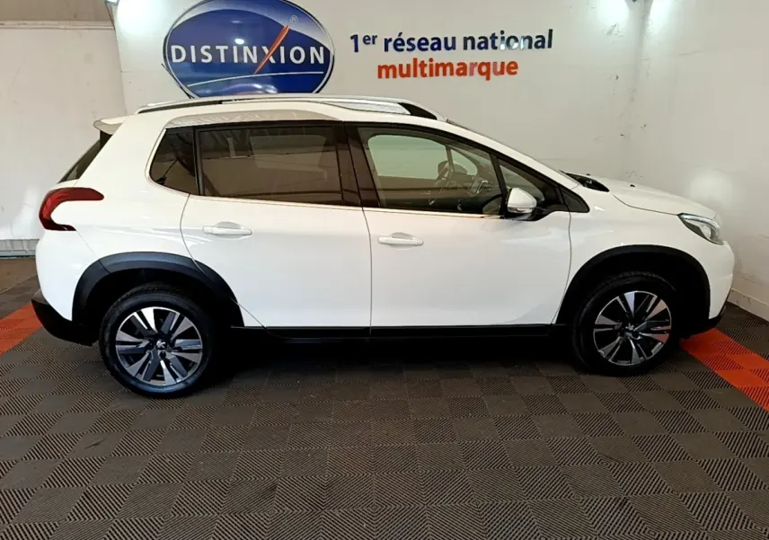 Vue de profil côté gauche d’un Peugeot 2008 blanc avec jantes alliage et barres de toit chromées en intérieur showroom.