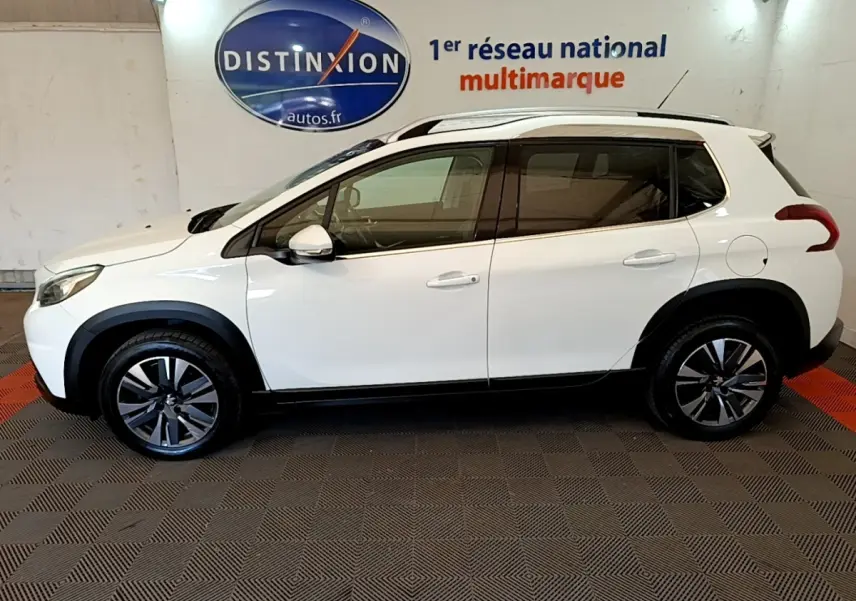 Vue latérale droite d'un Peugeot 2008 blanc avec jantes alliage et barres de toit chromées en intérieur showroom.