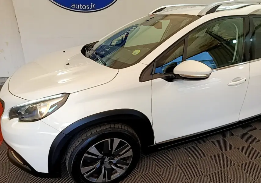 Vue 3/4 avant gauche d’un Peugeot 2008 blanc avec jantes alliage noires et barres de toit chromées.