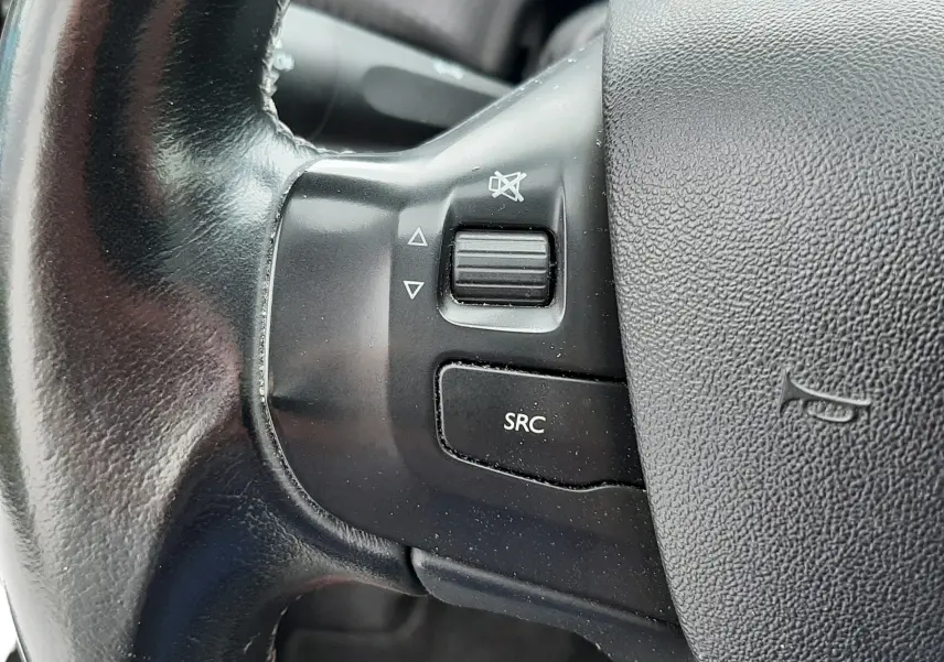 Gros plan sur les commandes gauche du volant noir du Peugeot 2008 blanc, avec bouton SRC et molette de volume.