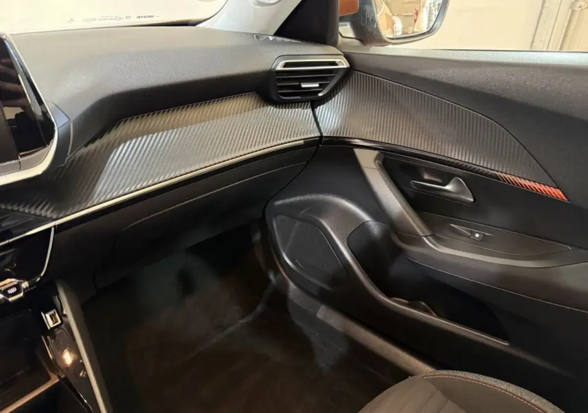 Vue intérieure côté passager du tableau de bord et porte avec insert décoratif effet carbone et surpiqûres orange.