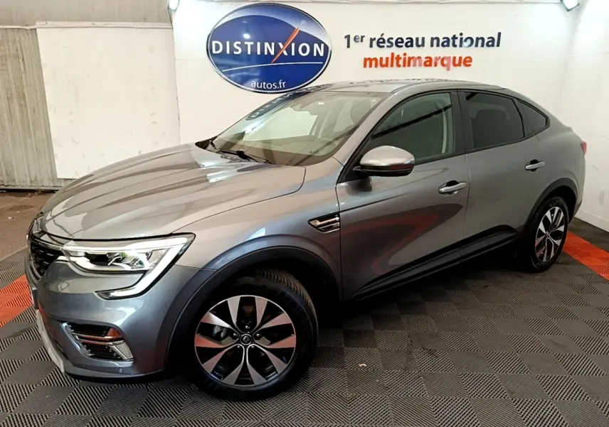 Vue 3/4 avant droit d’un Renault Arkana gris clair Evolution E-Tech full hybrid 145 en intérieur showroom.