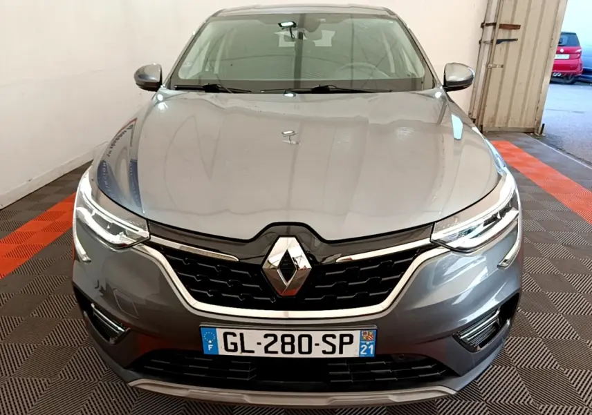 Vue frontale d'un Renault Arkana gris clair 2023 avec calandre noire et phares LED allumés dans un garage.