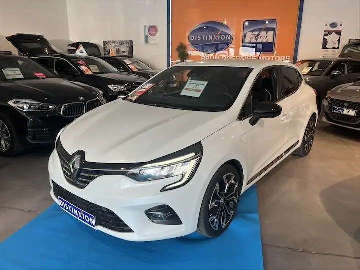 Renault Clio V blanc nacré métal vue 3/4 avant droit en showroom avec jantes noires et détails noirs contrastants.