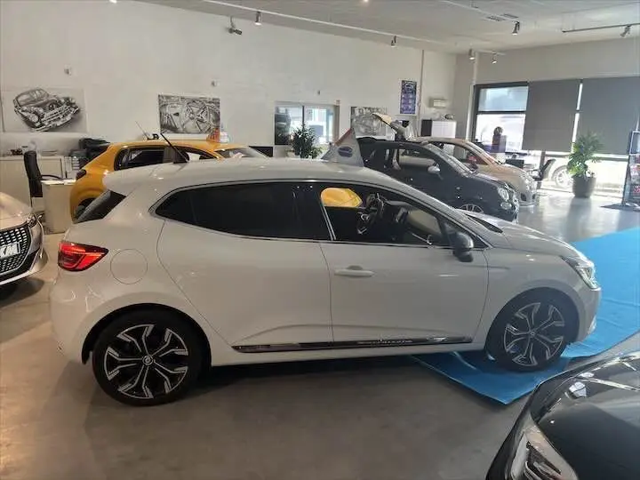 Profil côté gauche d'une Renault Clio V blanche nacrée métal en showroom, avec jantes noires et détails chromés visibles.