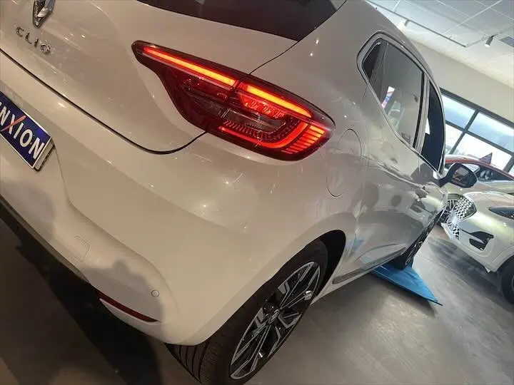 Vue 3/4 arrière droite d'une Renault Clio V blanche nacrée avec feux arrière allumés en intérieur showroom.
