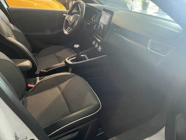 Intérieur de Renault Clio V blanc nacré, vue côté conducteur montrant sièges tissu noir et console centrale avec écran tactile.