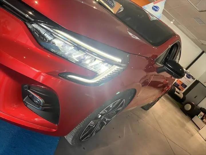 Vue 3/4 avant gauche d'une Renault Clio V rouge flamme métal avec phares LED allumés en intérieur showroom.