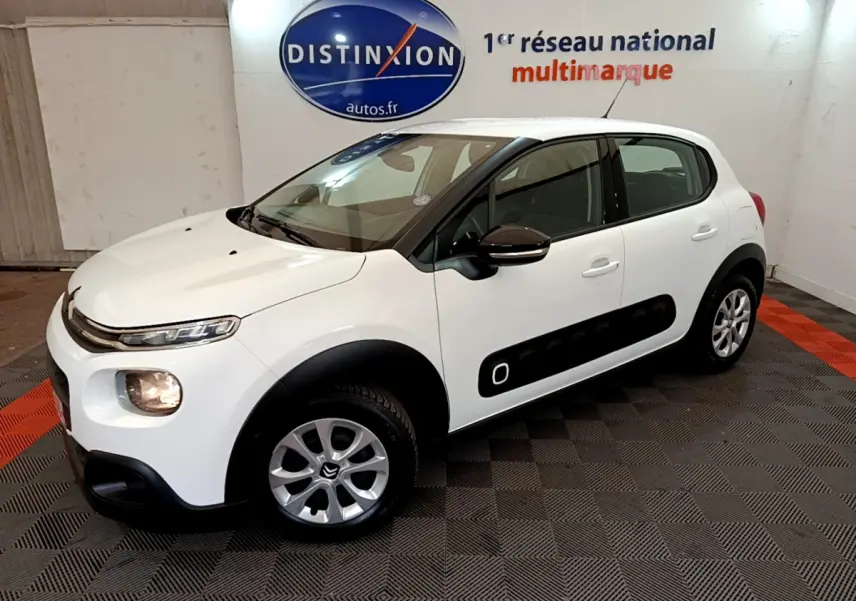 Citroën C3 blanche vue 3/4 avant droit, avec protections latérales noires et jantes alliage en intérieur showroom.