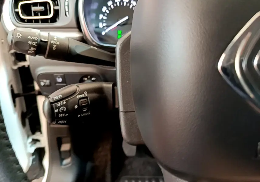 Gros plan sur le volant cuir et les commandes de régulateur de vitesse à gauche du tableau de bord d'une Citroën C3 blanche.